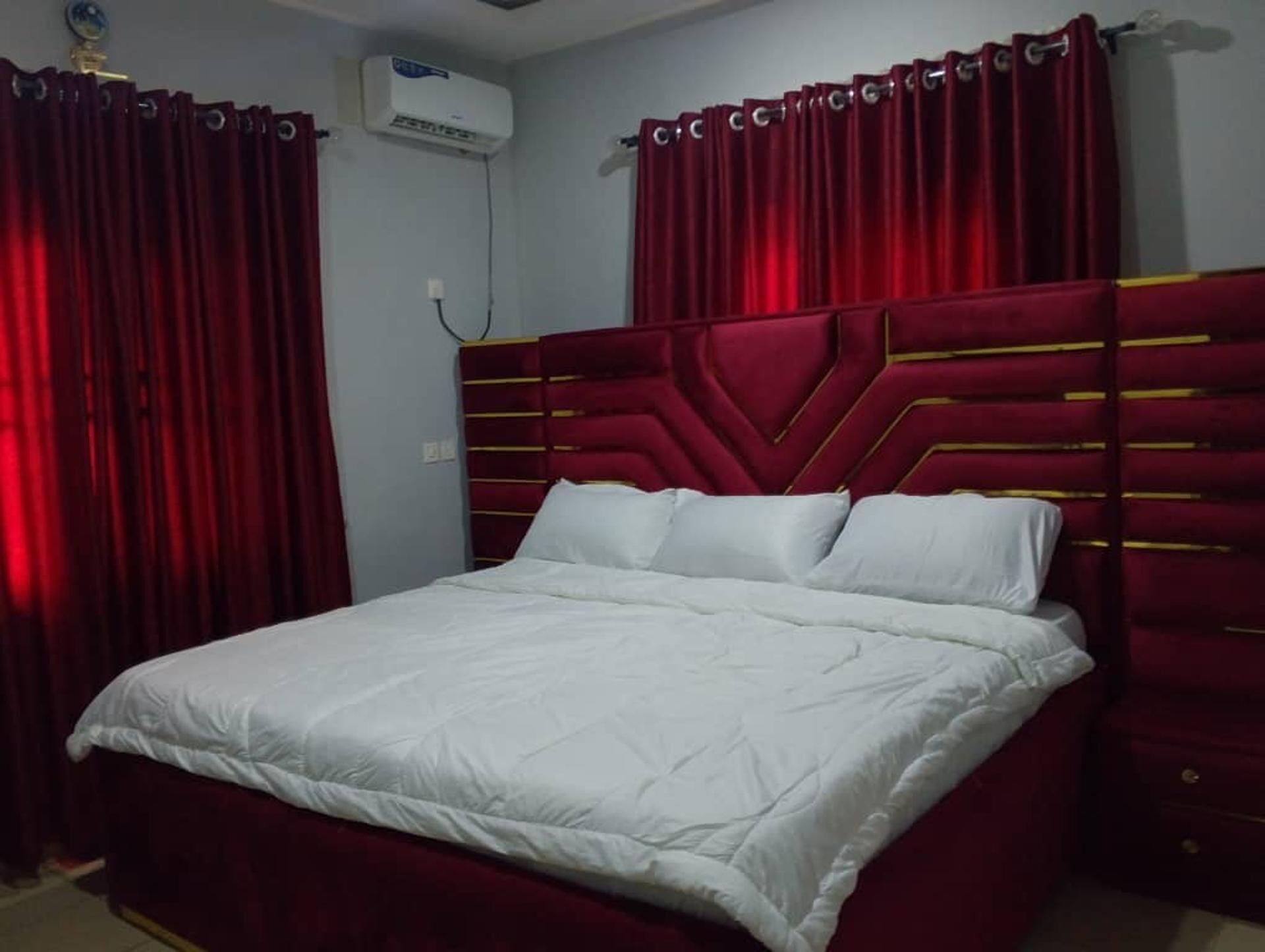 Un lit avec une couvre-lit blanche et des oreillers, une tête de lit rouge agrémentée de motifs dorés, et des rideaux rouges.