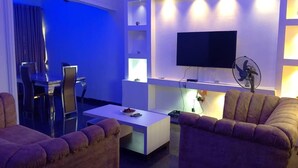 Egy modern nappali, amelyben található egy síkképernyős televízió, egy étkező asztallal és székekkel, valamint egy kanapé.