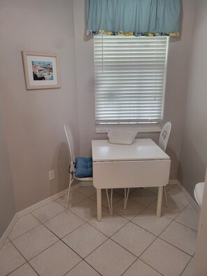 Een kleine kamer met een witte tafel, een blauwe stoel, een raam met zonwering, een lijst met een foto aan de muur, en een betegelde vloer.