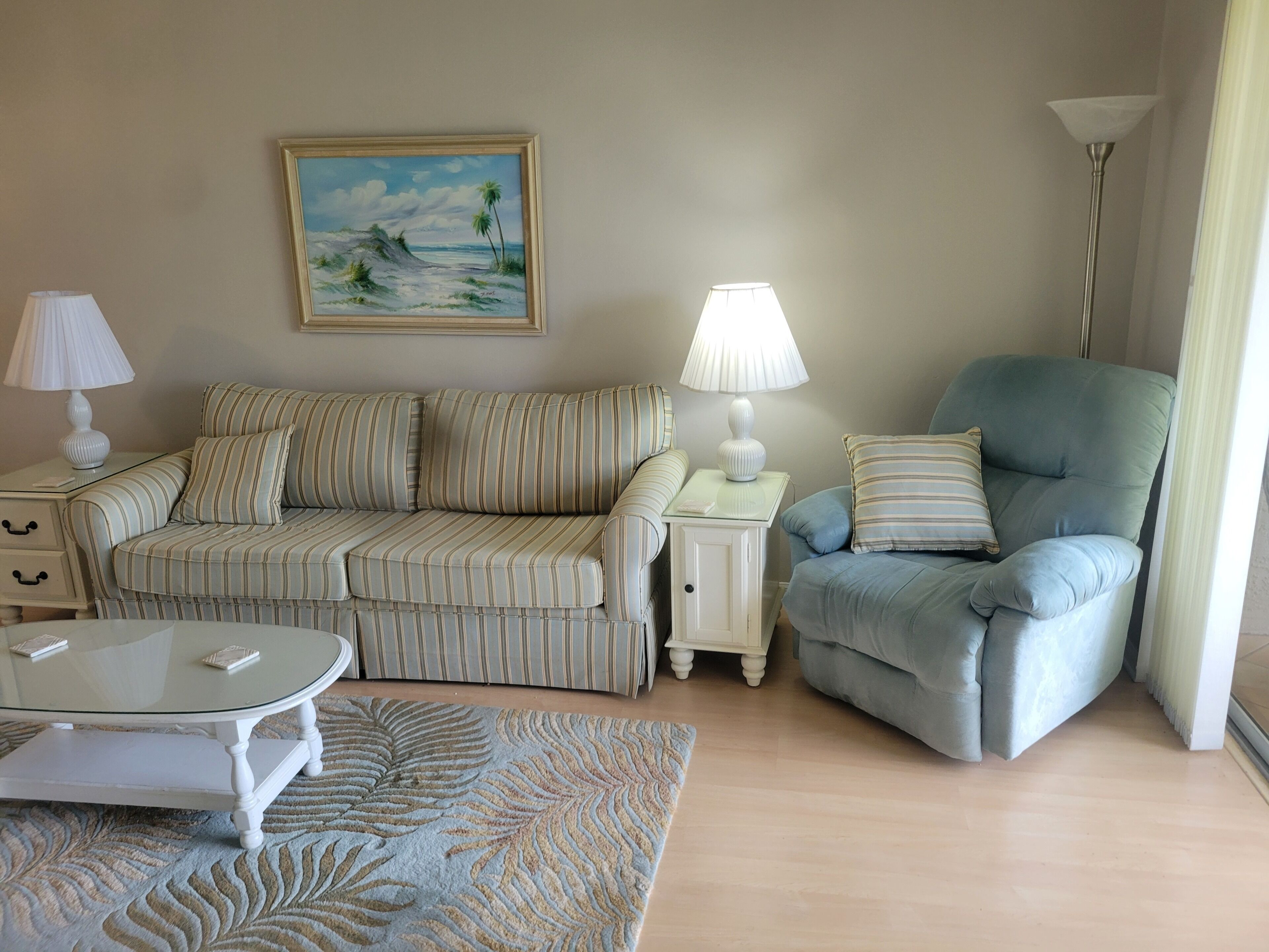 Un salon avec un canapé à rayures, un fauteuil bleu, une table basse en verre, un tableau représentant une scène de plage et un lampadaire.
