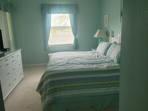 Een slaapkamer met een bed, een dressoir, een raam met gordijnen en een lamp op een nachtkastje.