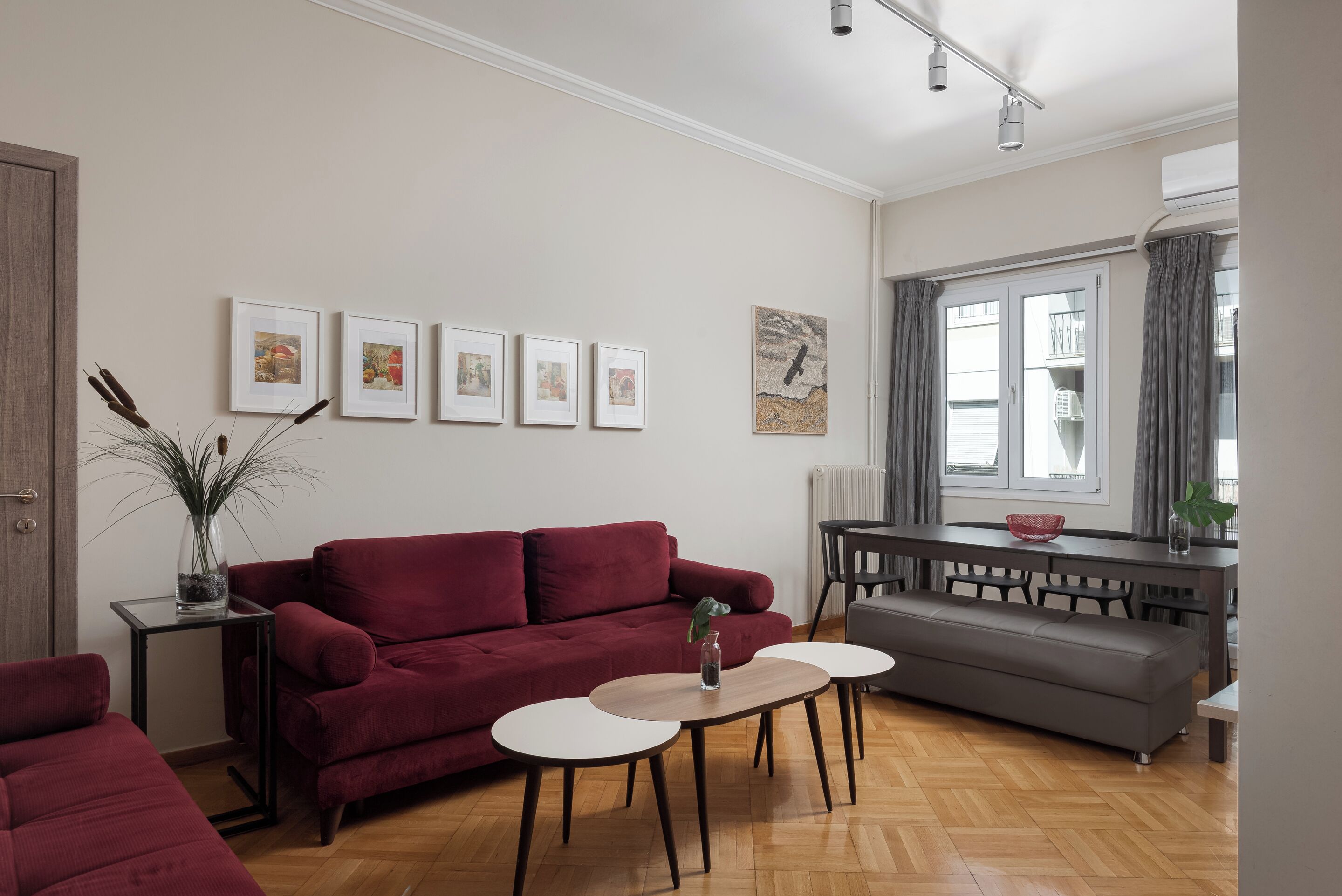 A modern living room with a red sofa, two white round tables, and a grey sofa. There are framed pictures on the wall, a plant on a side table, and a window with curtains.