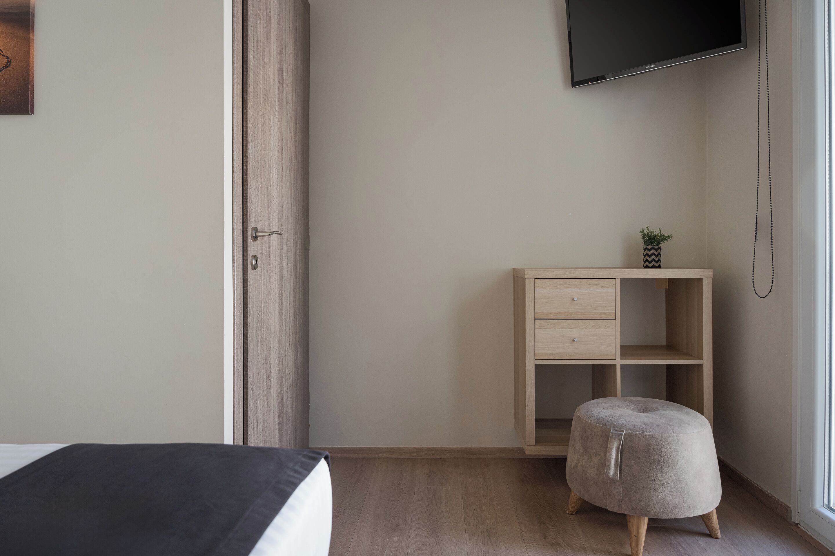 A bedroom with a bed, a wooden nightstand, a small stool, a TV mounted on the wall, and a window with a view.