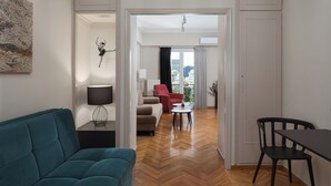 A modern living room with a teal sofa, a dining table with chairs, and a view of the city through large windows. - Athenian Heritage Residences (Athens)
