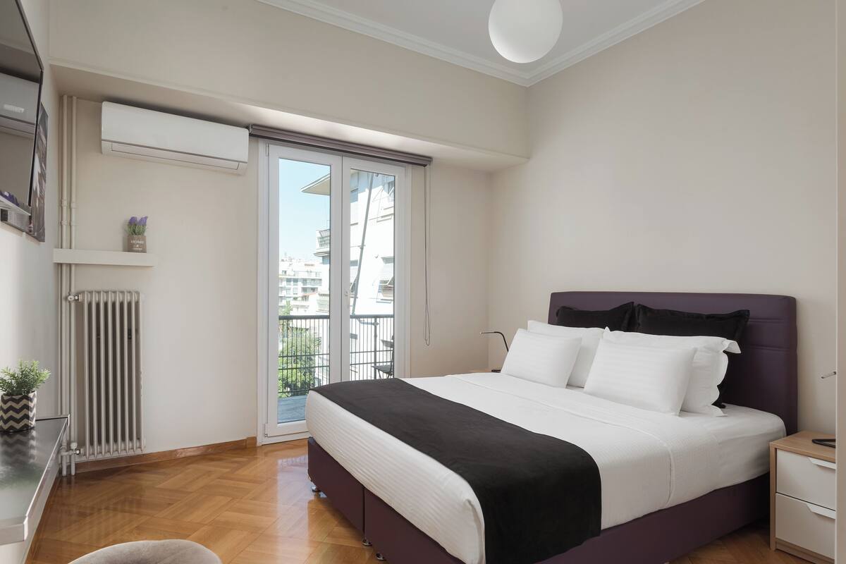 Une chambre à coucher comprenant un lit, une table de chevet, une commode, un radiateur et une porte-fenêtre coulissante donnant sur un balcon.