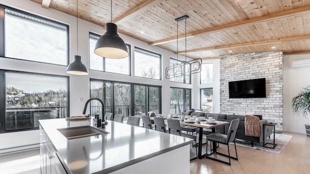 A modern kitchen with a large island, dining area with a sofa and table, and a TV mounted on a brick wall. - Spa & Mountain View | Le Eastmain View (Saint-Côme)