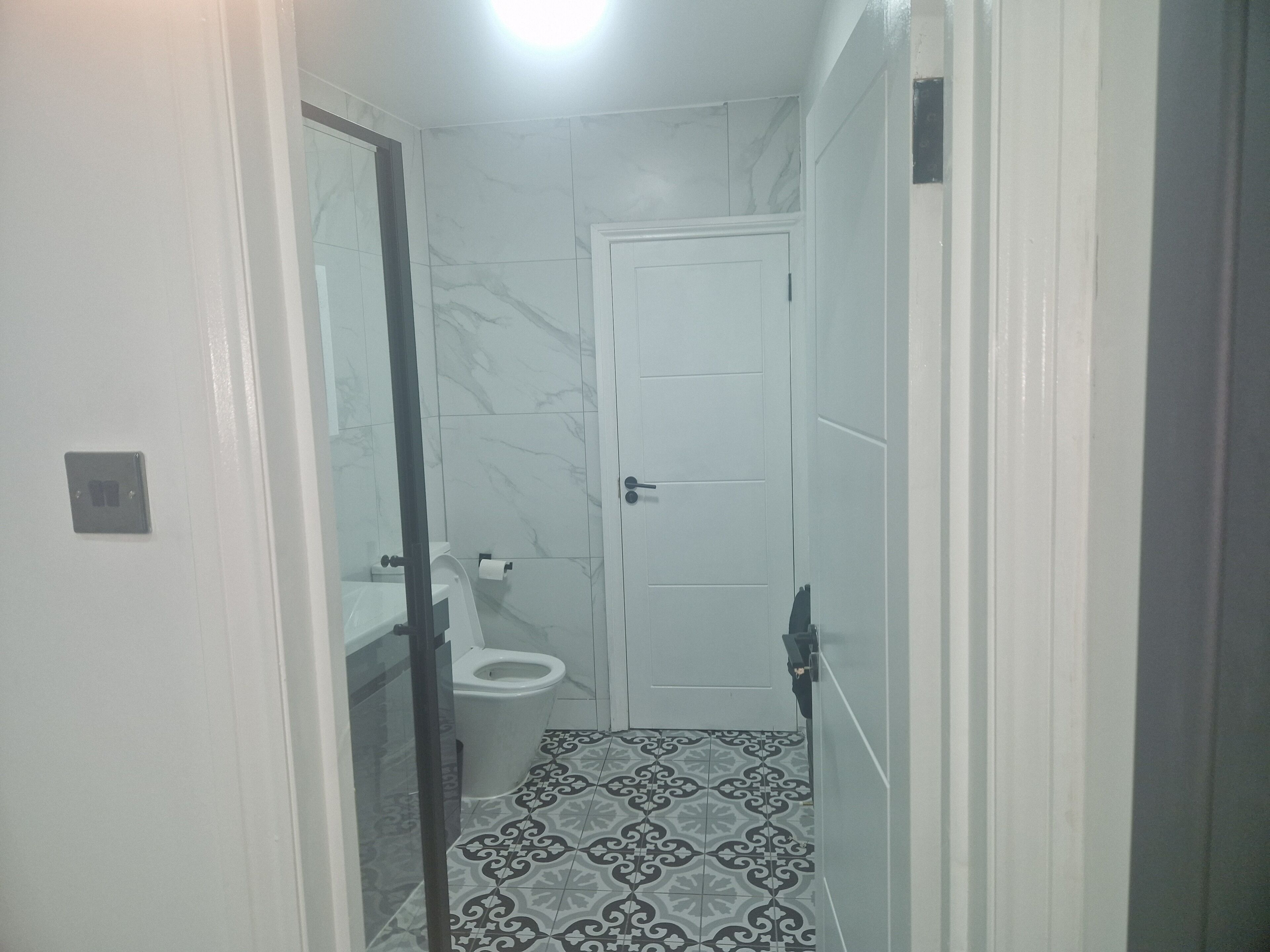 Une petite salle de bain comprenant des toilettes, un lavabo et un miroir. Le sol est recouvert de carreaux à motifs, et les murs sont recouverts de carreaux imitant le marbre.