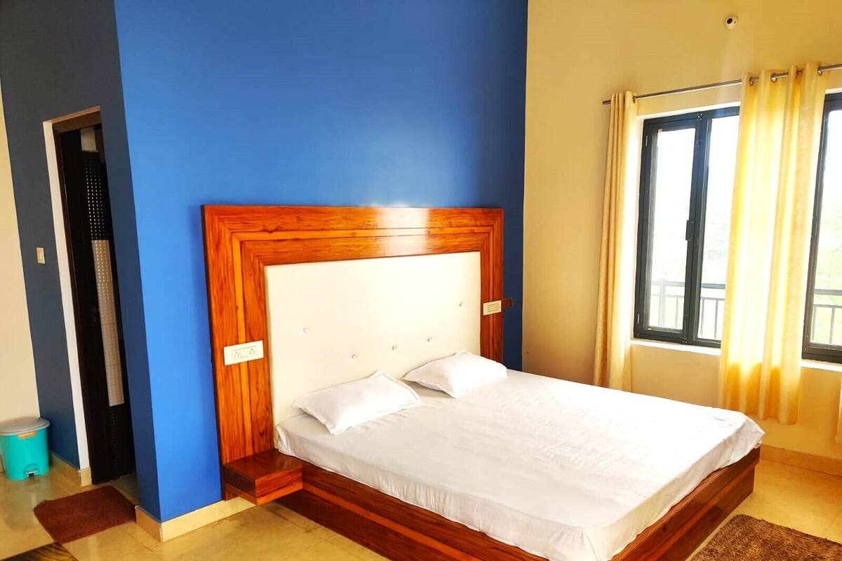 A bedroom with a wooden headboard, a bed with white bedding, a window with curtains, and a door with a towel rack.
