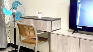 A room with a desk, chair, standing fan, TV, and a small shelf with a decorative item. - Napper's Secret Peaceful Place (Kampala)