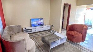 A living room with a sofa, a red armchair, a coffee table, and a TV on a white cabinet. - Napper's Secret Peaceful Place (Kampala)