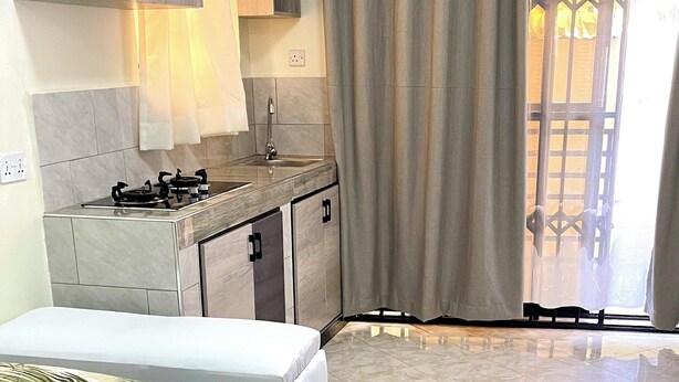 A bedroom with a bed, a nightstand, a window with curtains, and a bathroom with a sink and mirror. - Napper's Secret Peaceful Place (Kampala)