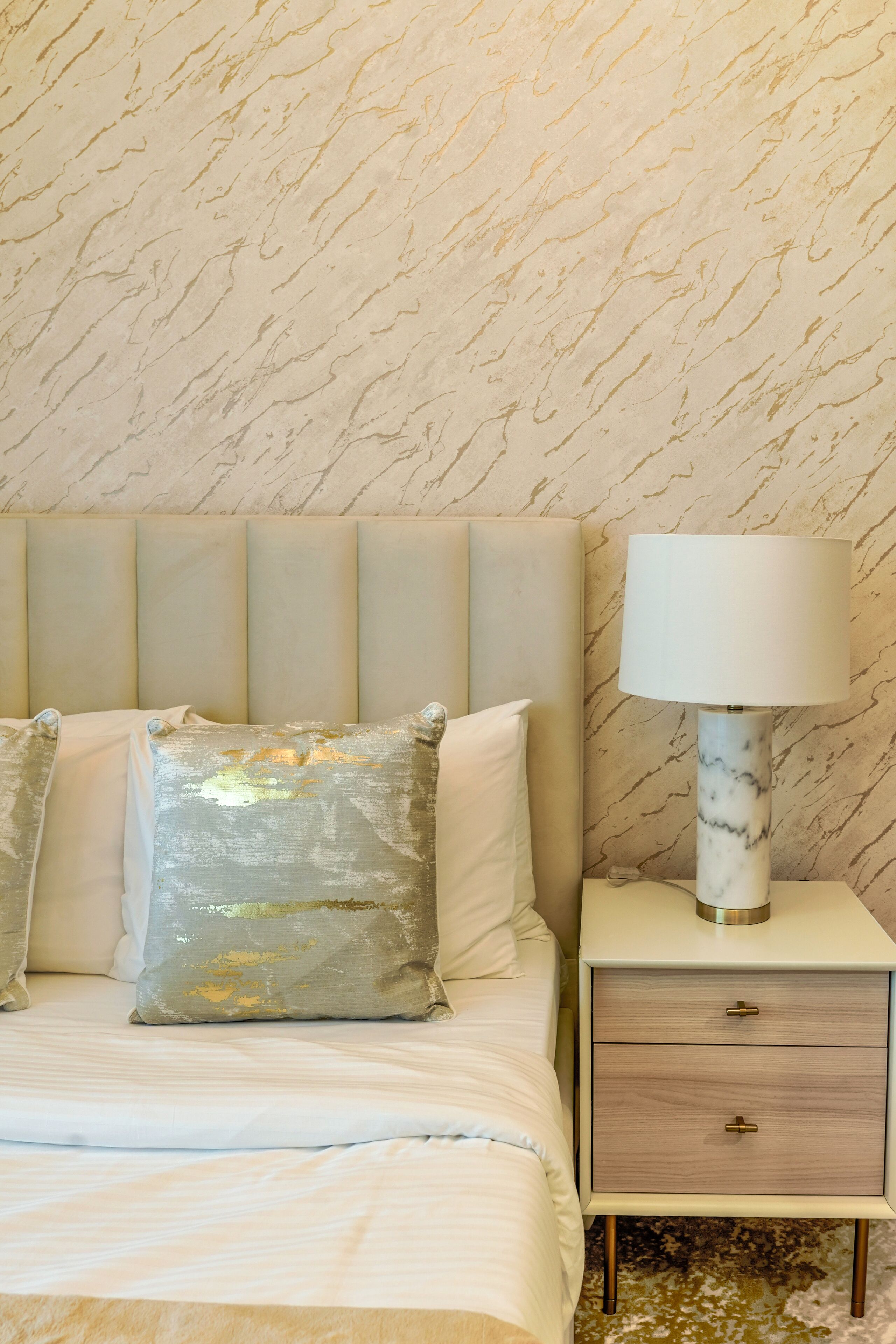 Una cama bien hecha con cabecera, dos almohadas, una mesita de noche con lámpara y una pared con diseño de mármol.