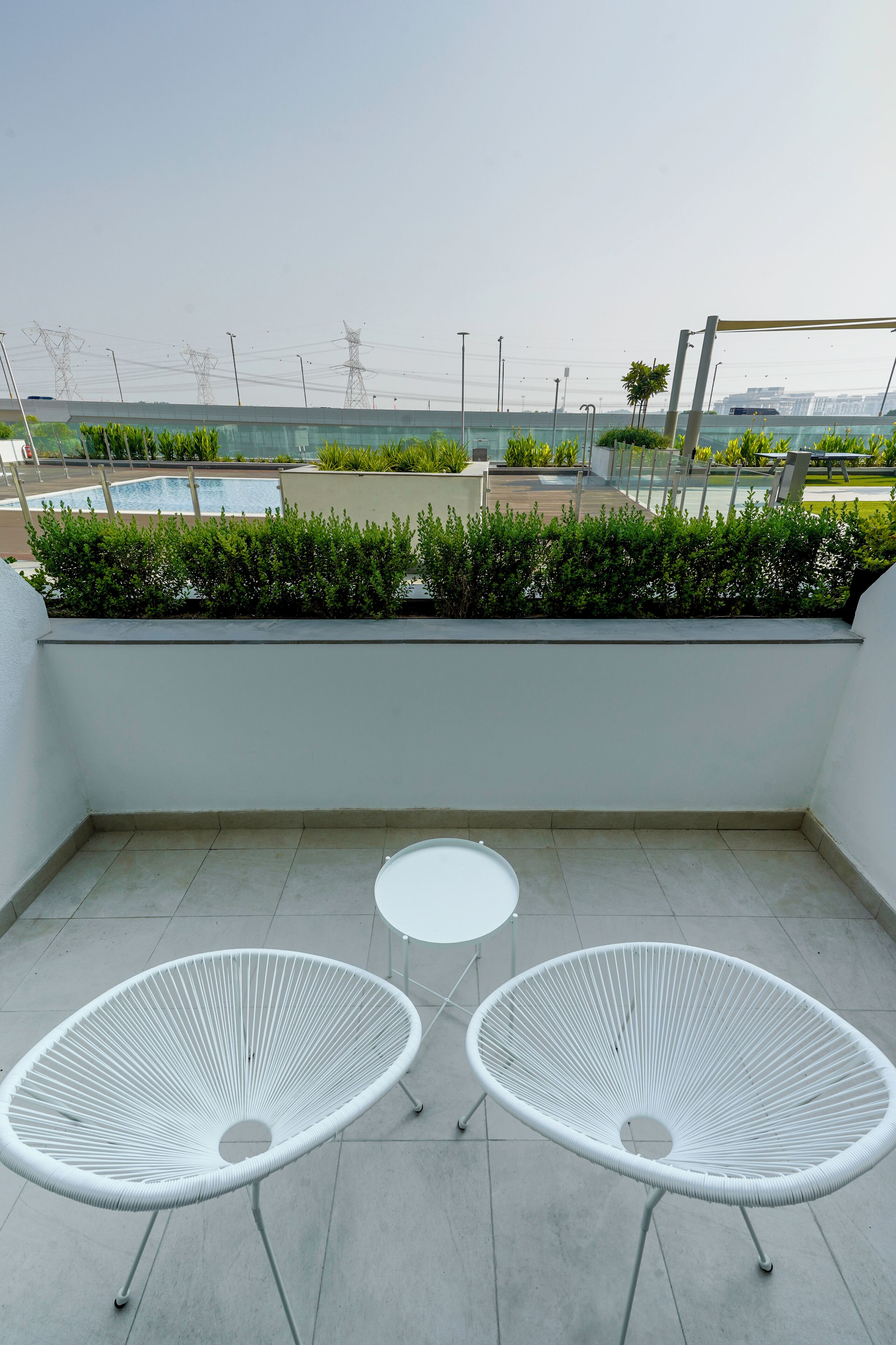 A rooftop terrace with two white wire chairs, a small round table, and a view of a swimming pool and greenery.