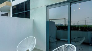 A modern outdoor seating area with white chairs and a small table, a glass door leading to a balcony, and a view of the cityscape. - Nasma Holiday Homes - Azizi Riviera 46 (Dubai)