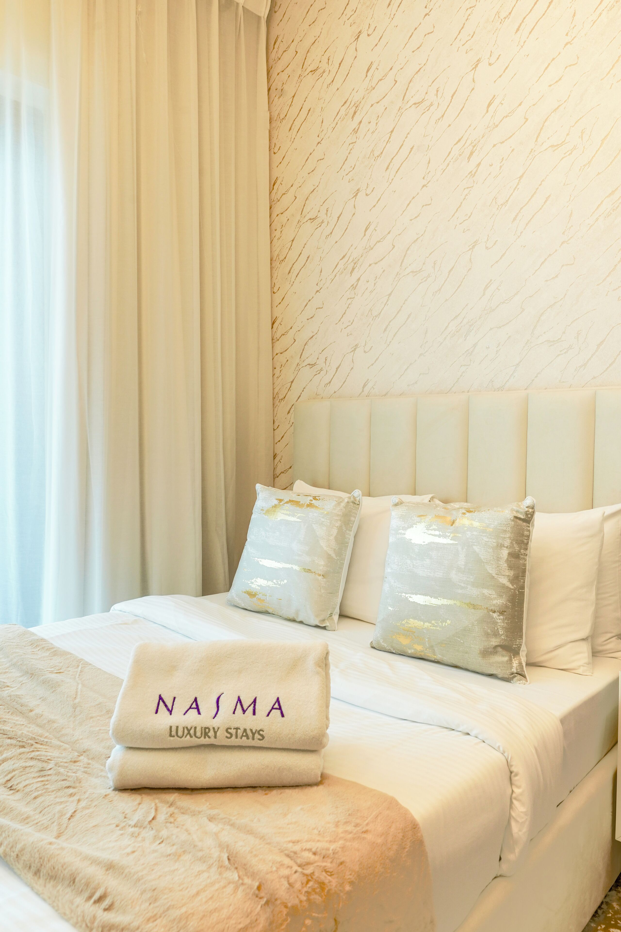 A neatly made bed with white linens, two decorative pillows, and a folded towel with "NASMA LUXURY STAYS" text.