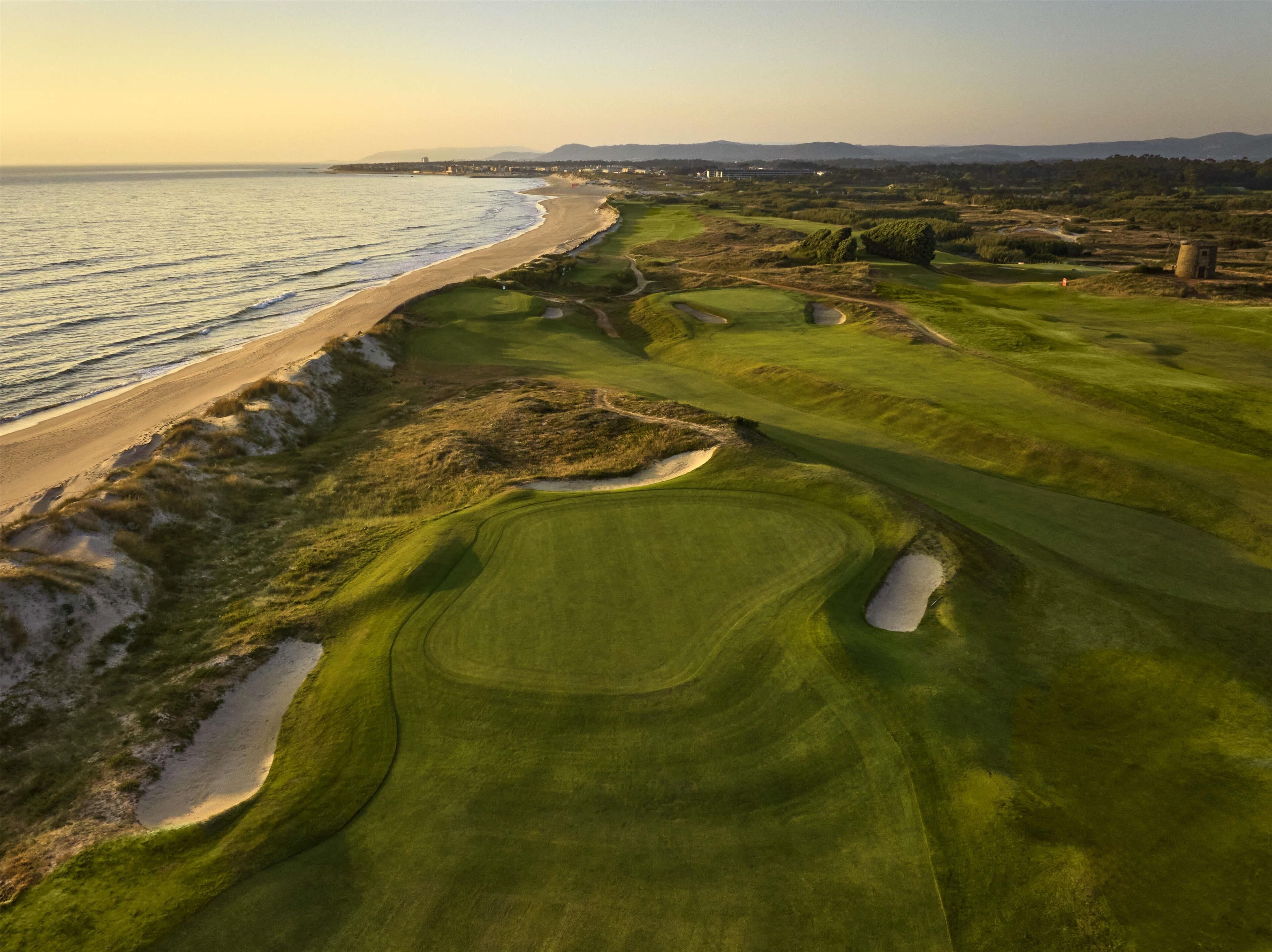 Un campo de golf con vista al mar, búnkeres de arena y una cadena montañosa distante.