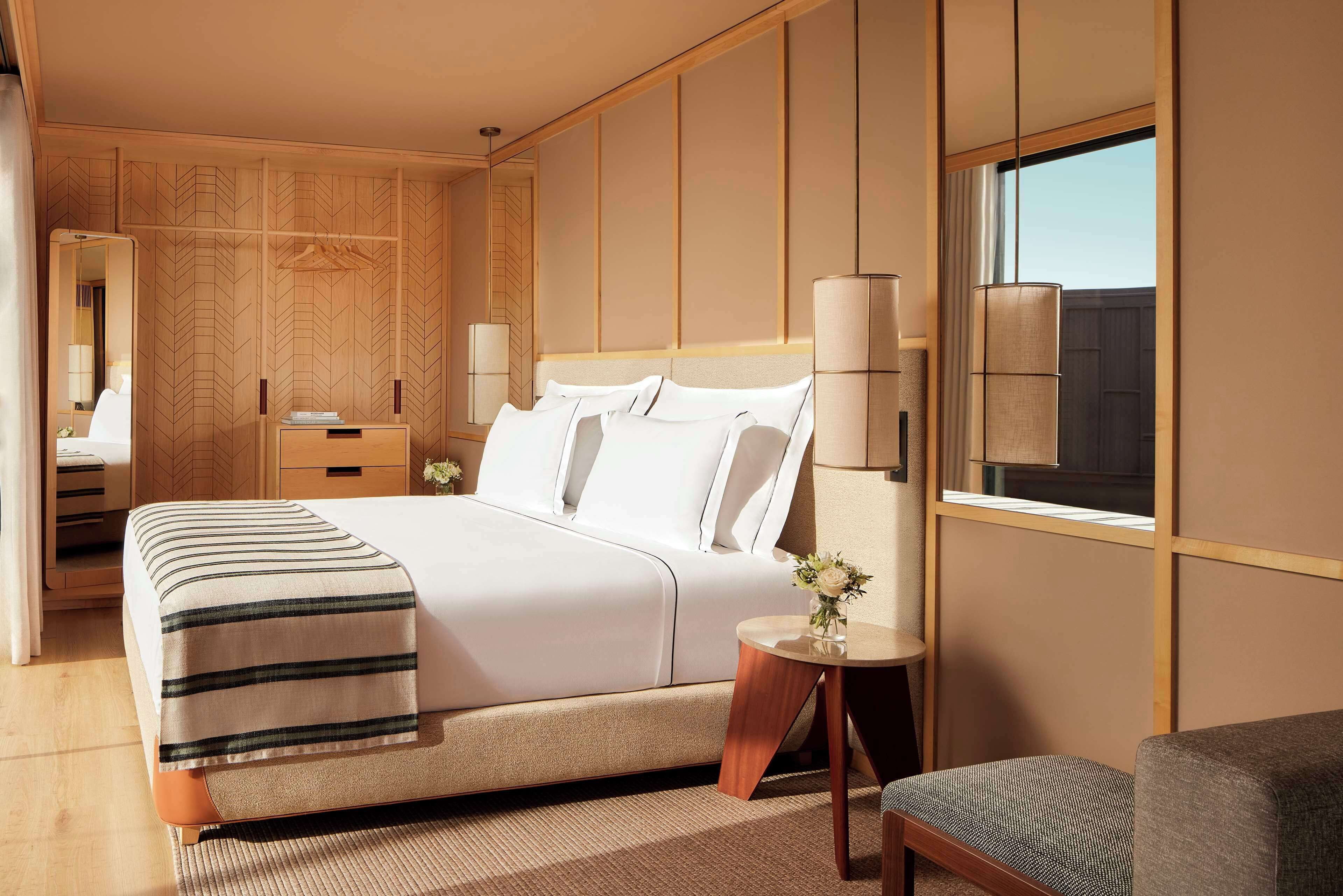 Una habitación de hotel moderna con una cama grande, mesitas de noche, una pequeña mesa redonda con un jarrón de flores y vistas al exterior a través de una ventana.