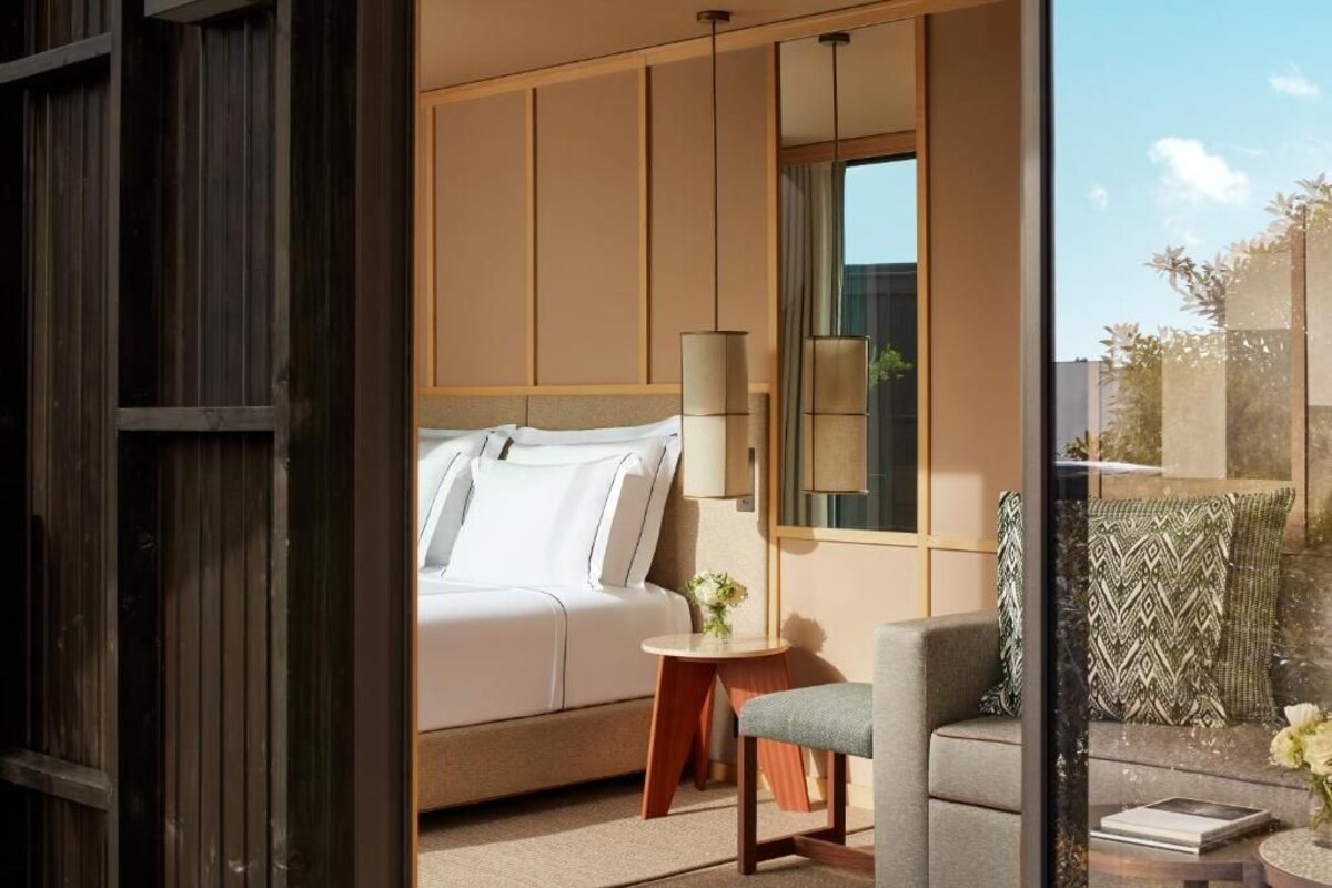 A bedroom with a bed, bedside table, lamp, and a view of the outside.
