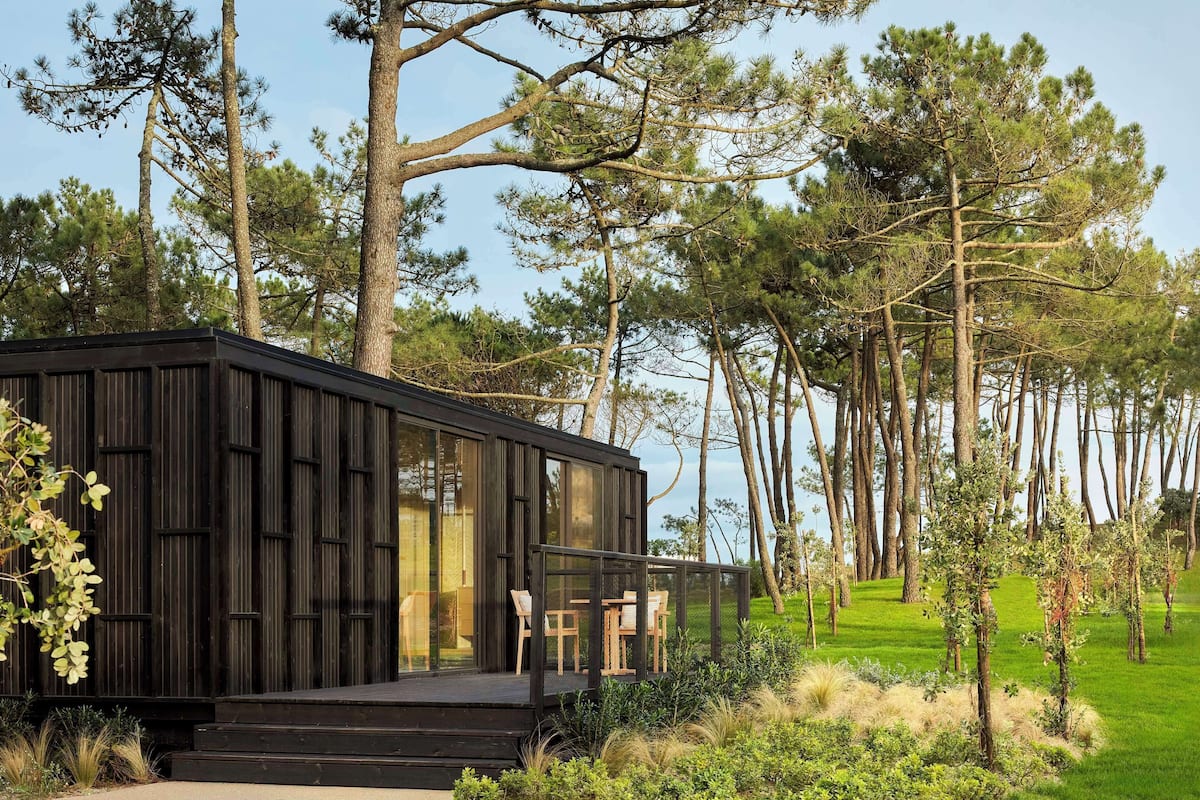 Une cabane moderne à la façade vitrée, entourée d’un paysage naturel composé de grands arbres et de prairies.