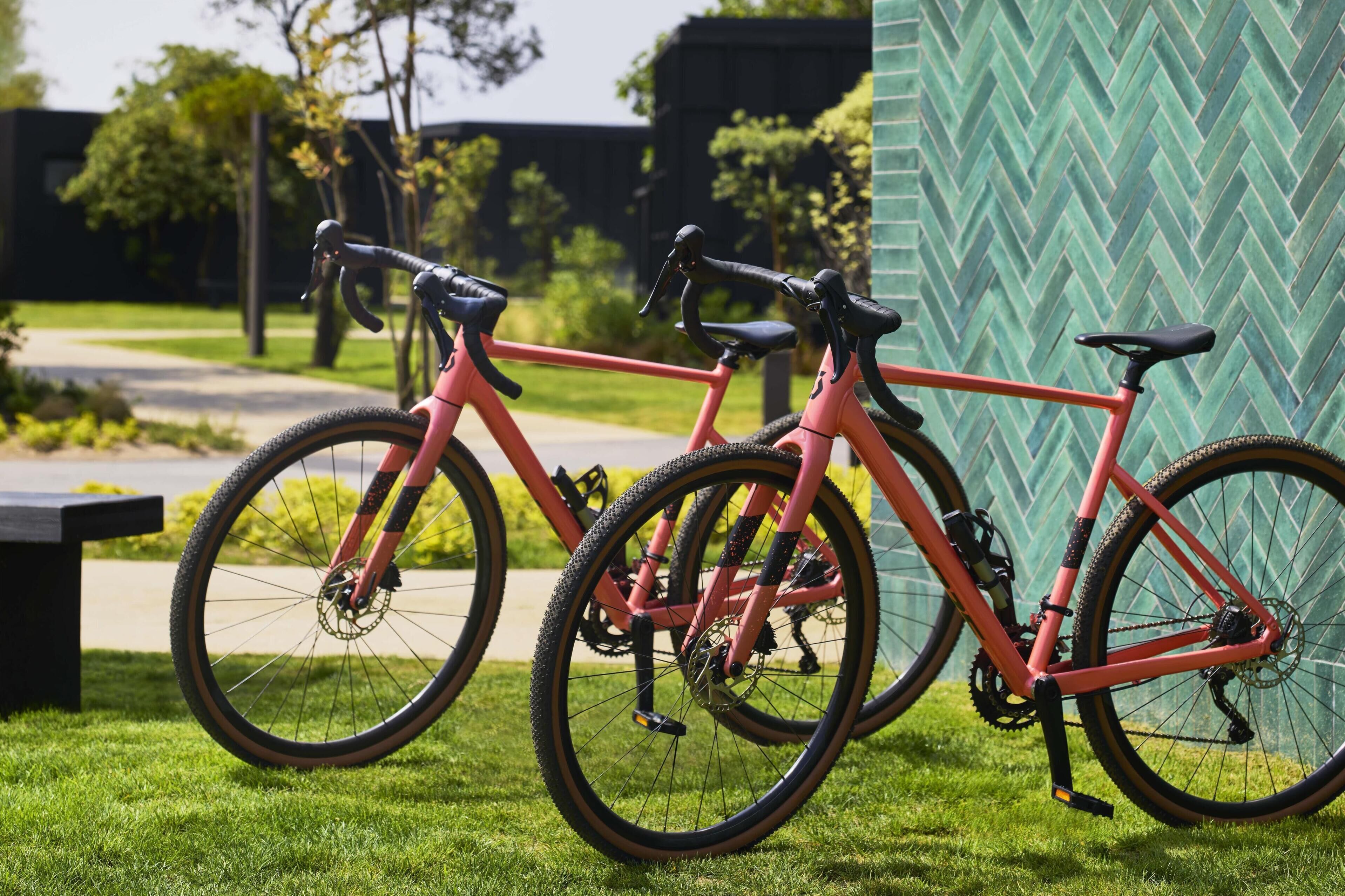 Two bicycles parked on a grassy area with a green wall and a building in the background.