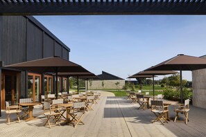 Un patio al aire libre con mesas y sillas de madera, grandes sombrillas y vistas a un jardín ajardinado.