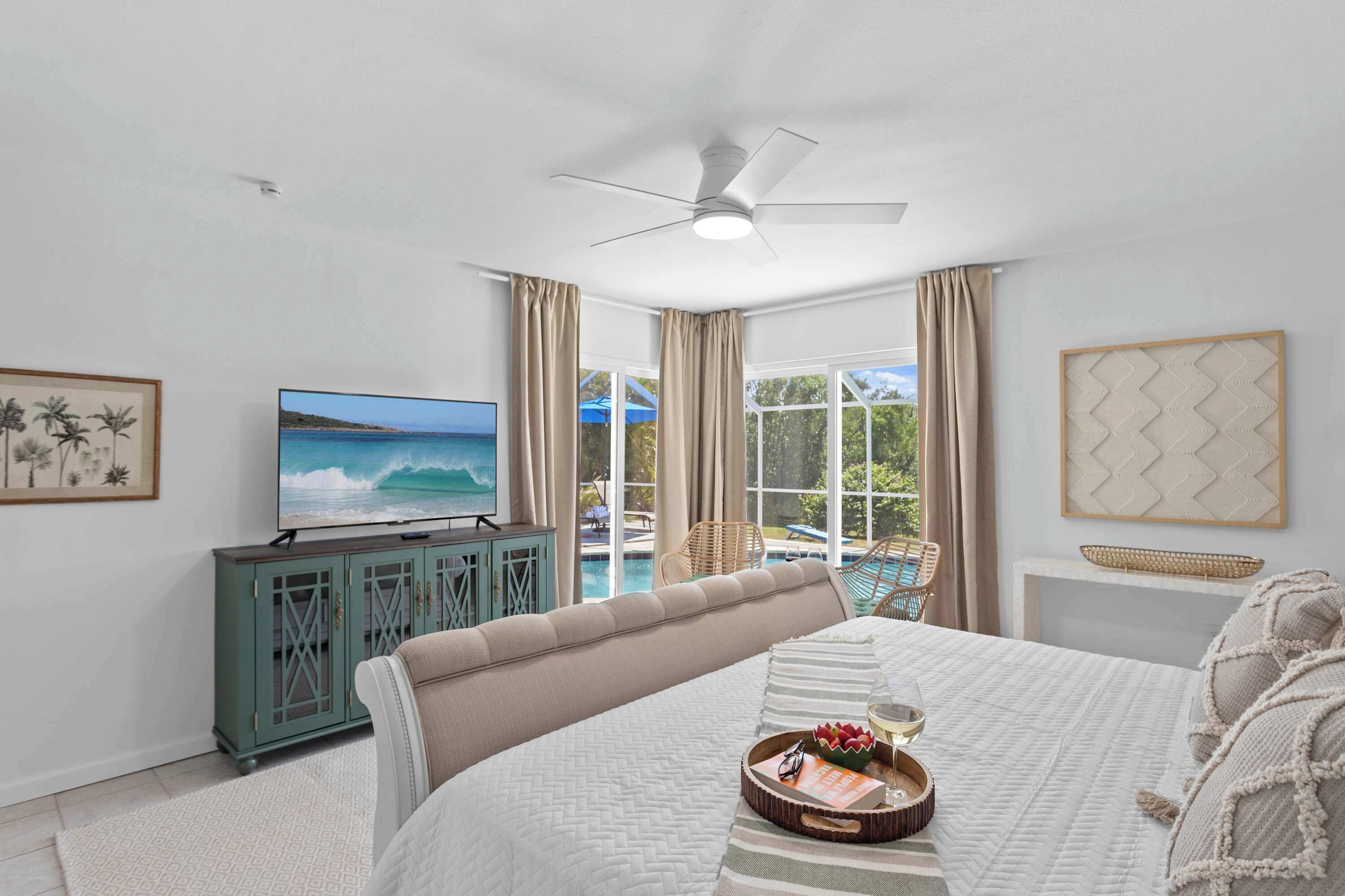 A bedroom with a bed, a TV, a ceiling fan, a window with curtains, a view of a pool, and a framed picture on the wall.