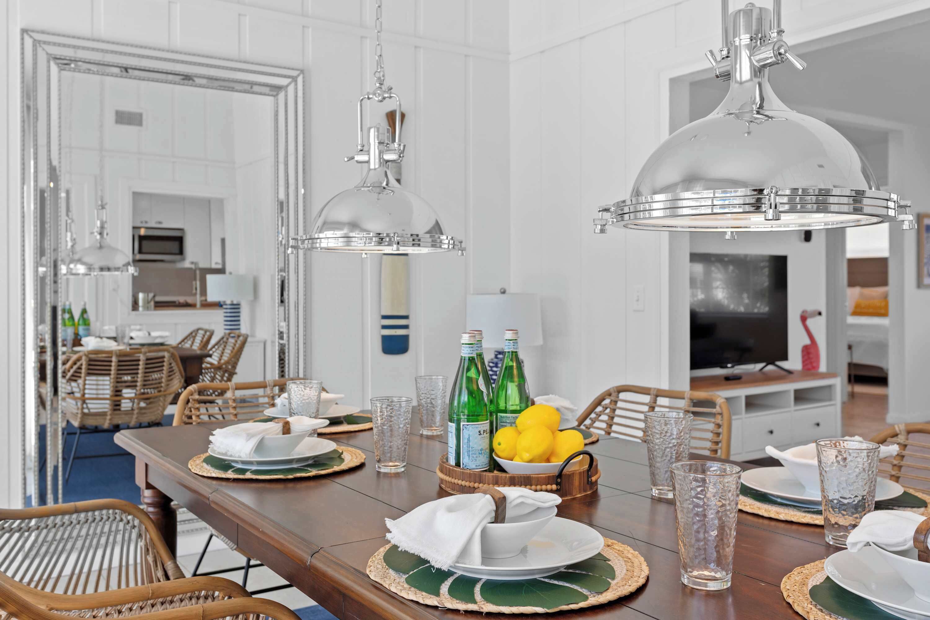 A dining area with a wooden table set for six, surrounded by wicker chairs. There are two pendant lights above the table, and a television is visible in the background.