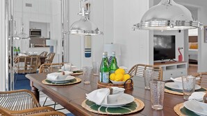 A dining area with a wooden table set for six, surrounded by wicker chairs. There are two pendant lights above the table, and a television is visible in the background. - Heated Pool, Bay Views & Kayak Launch — 6 Guests (Bradenton)