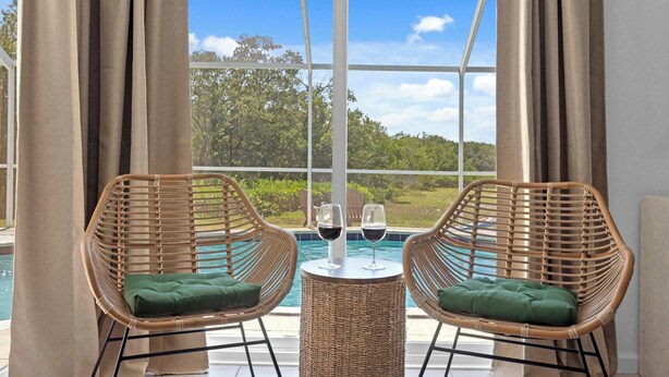 Two wicker chairs with green cushions, a small round table with two wine glasses, and a view of a swimming pool and greenery through large windows. - Heated Pool, Bay Views & Kayak Launch — 6 Guests (Bradenton)