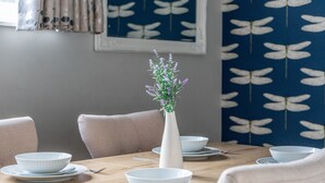 Una mesa de comedor dispuesta con cuencos y platos blancos, un florero blanco con flores y una pared azul con un diseño blanco.