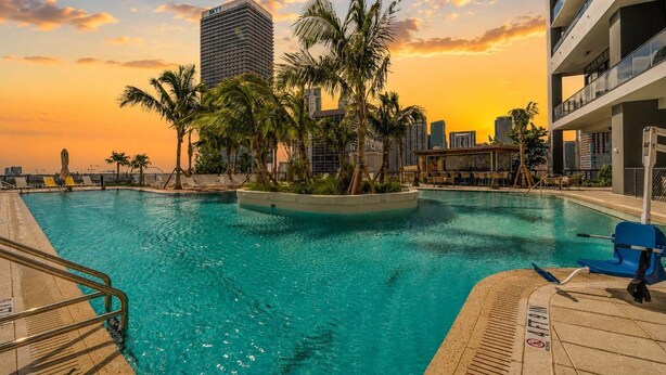 Una piscina con palmeras, el perfil de una ciudad y un atardecer.