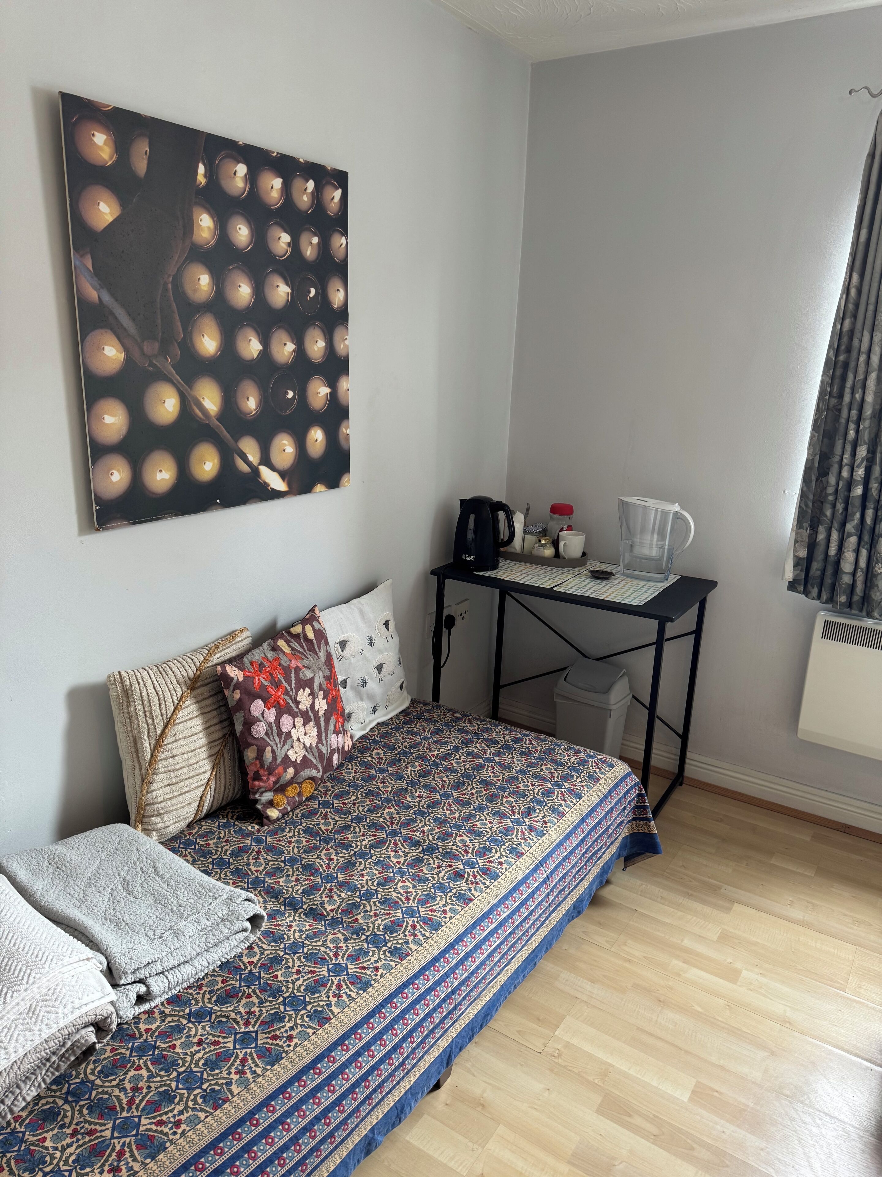 A bedroom with a bed, a desk, a kettle, a cup, and a radiator.