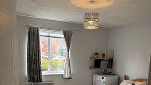 A bedroom with a bed, a bench, a radiator, a window with curtains, a hanging light fixture, and a microwave. - Spacious Tidy Room in Cardiff City (Cardiff)