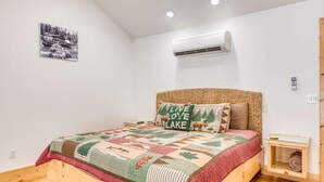 Une chambre avec un lit en bois, une courtepointe ornée de motifs représentant la forêt et la faune, un climatiseur fixé au mur et une petite table de chevet avec une lampe.