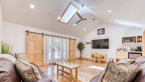 Un salon spacieux avec un grand canapé en cuir marron, une table basse en bois et un ventilateur de plafond. Une porte-fenêtre coulissante donne accès à un balcon, et un téléviseur est fixé au mur.