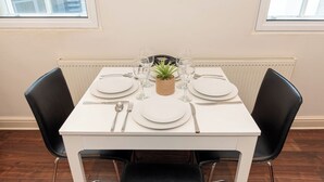 Mesa de jantar posta para quatro com pratos brancos, talheres e copos transparentes. No centro, uma pequena cesta trançada com uma planta verde. Duas cadeiras pretas estão posicionadas de cada lado da mesa.
