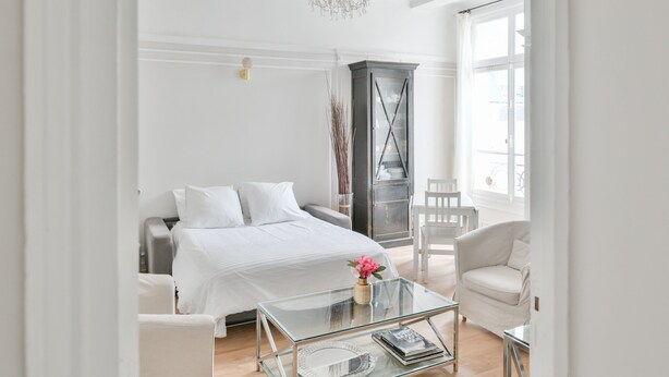 Un dormitorio moderno con una cama, una mesita de vidrio, un sillón blanco y un aparador alto.