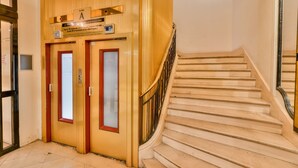 Ascensor de hotel con reloj digital, un cartel que indica el piso y una escalera con barandilla.