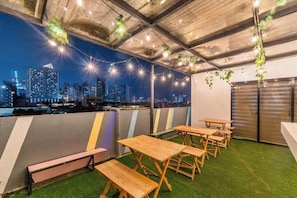 An outdoor seating area with tables and benches, a city skyline view, and string lights. - Redenza Suites Makati (Makati)