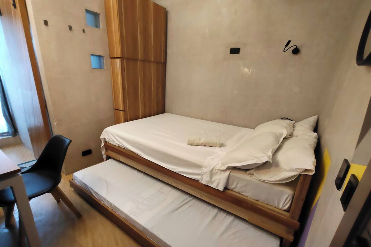 A single bed with a wooden frame and white bedding, a black chair, and a small desk in a room with a plain wall and a wooden cabinet.