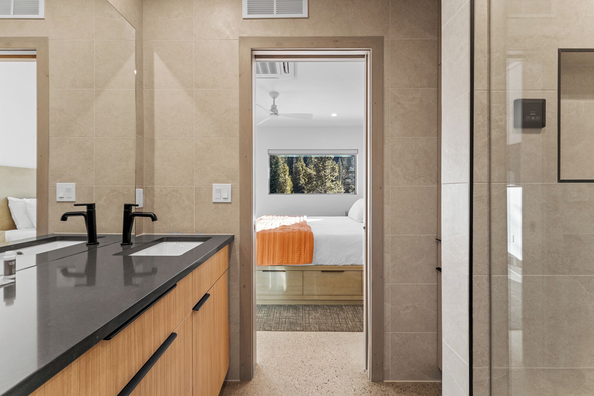 Ein modernes Badezimmer mit einem Doppelwaschbecken, einem Spiegel und Blick auf ein Schlafzimmer mit Bett und Fenster.