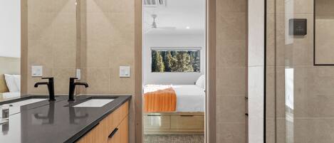 Ein modernes Badezimmer mit einem Doppelwaschbecken, einem Spiegel und Blick auf ein Schlafzimmer mit Bett und Fenster.