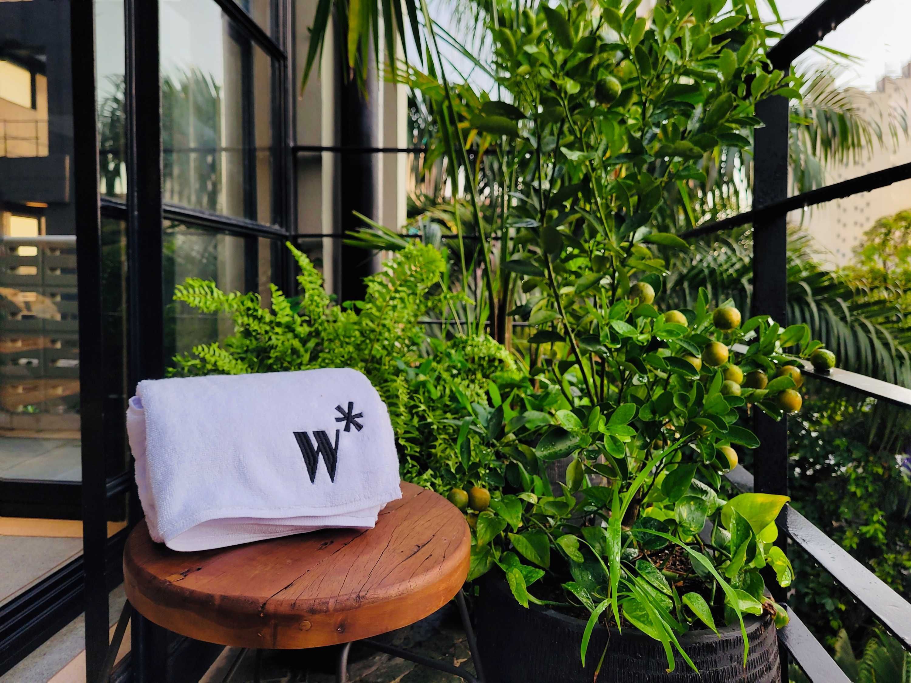 Een witte handdoek met een zwarte letter 'W' op een houten kruk op een balkon met plantenbakken en een gebouw op de achtergrond.