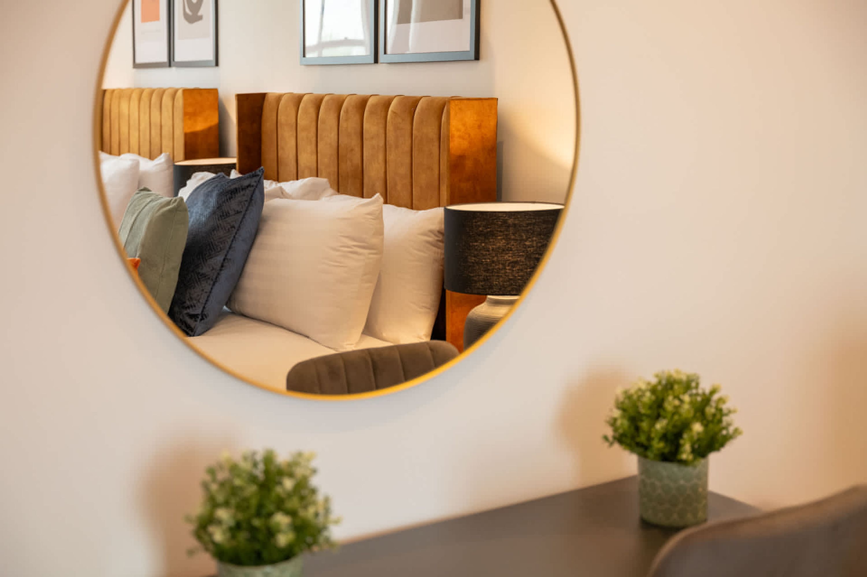 A circular mirror reflecting a bed with a wooden headboard, a bedside lamp, and two small potted plants on a table.