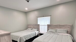 A bedroom with two beds, a window with blinds, a ceiling light, and a wooden floor.
