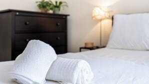 A neatly made bed with a white towel, a dark wooden dresser, and a lamp with a white shade. - Urban City View Apartment (Lagos)