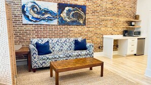 Un salon avec un mur en briques, un canapé à motifs, une table basse en bois et un coin cuisine équipé d’un four à micro-ondes.
