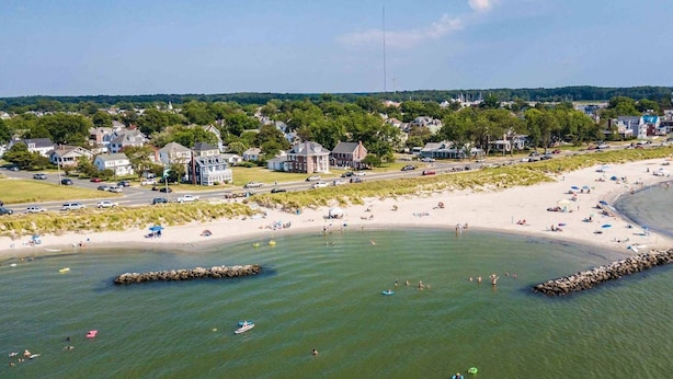 Une vue aérienne d’une plage, avec des maisons en arrière-plan, un rivage sablonneux et des eaux calmes.