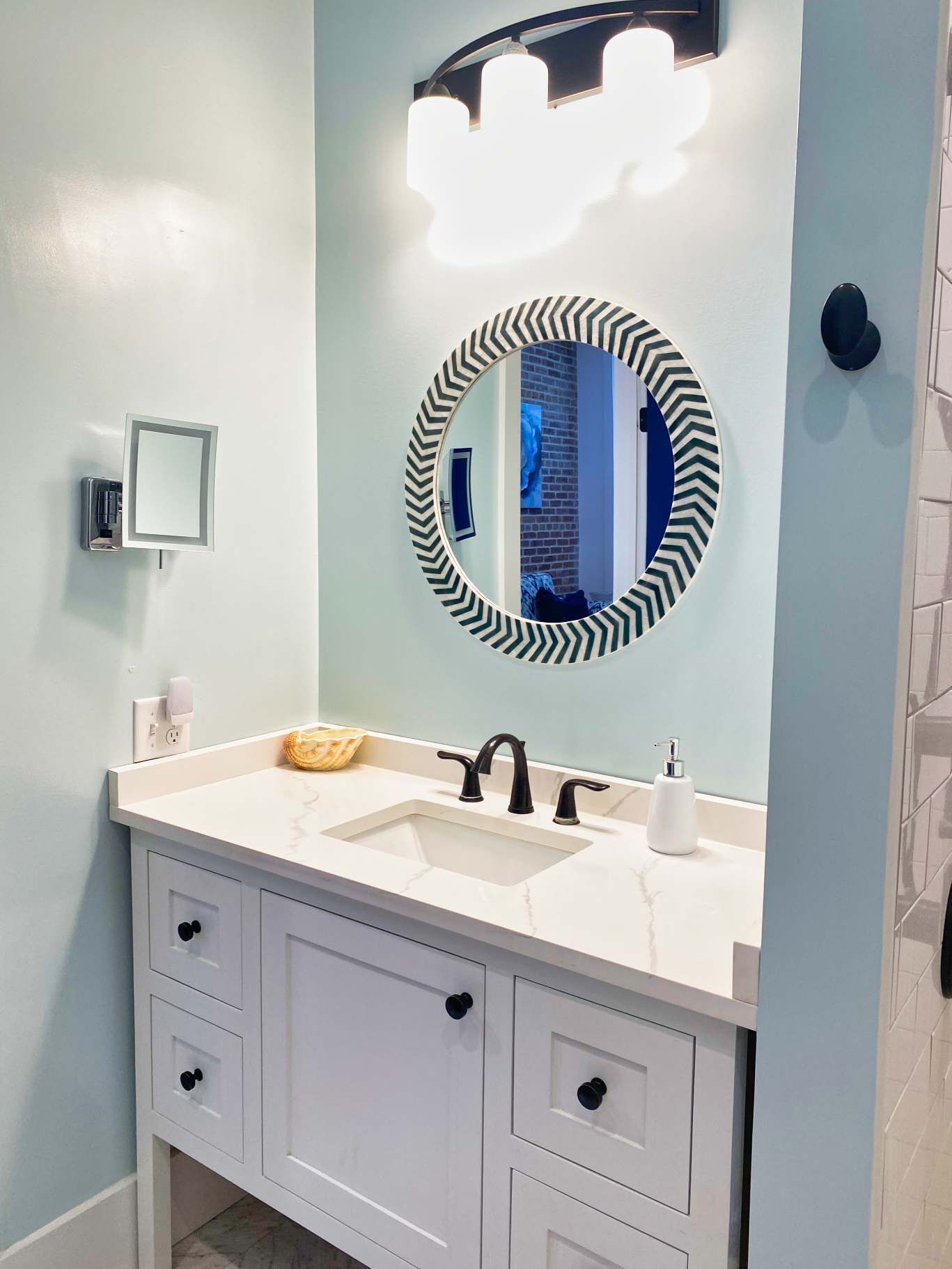 A bathroom with a white vanity, two sinks, a mirror with a black and white chevron frame, and a wall-mounted light fixture.
