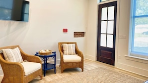 Une pièce comprenant deux fauteuils en osier, une petite table bleue, un tapis tissé, un téléviseur fixé au mur et une porte surmontée d’une fenêtre.
